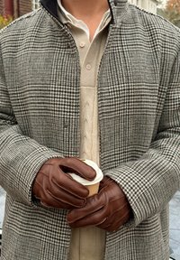 Brune skinnhansker holder en kaffekopp. En frakk med hundetannmønster i grått og svart ligger over en kremfarget kabelstrikket genser.