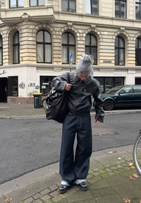 Zwarte leren jas met een ritszak, losse donkere denim jeans, grijze beanie en zwarte tas; staande op straten met kinderkopjes.