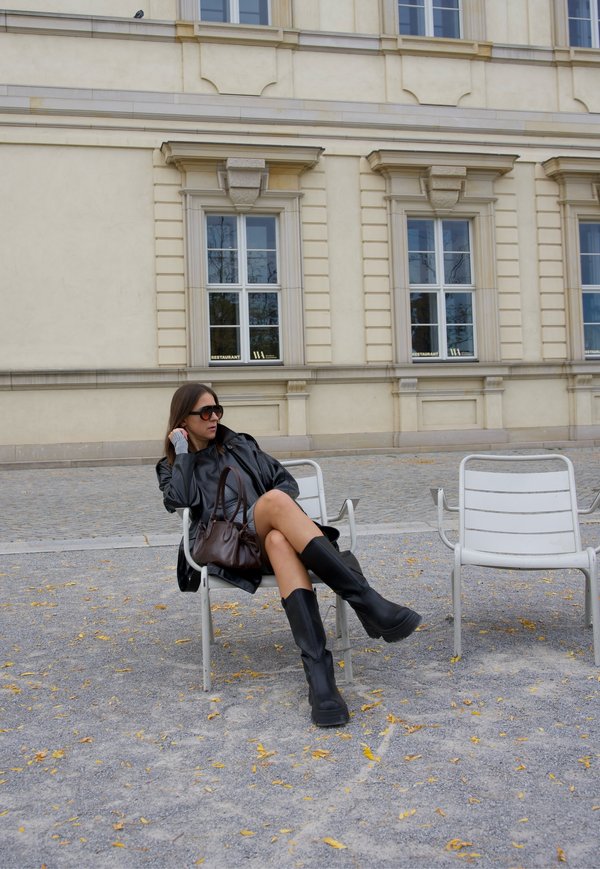 Femme en manteau noir, bottes et lunettes de soleil assise sur une chaise blanche à l'extérieur près d'un bâtiment beige avec des fenêtres étiquetées "Restaurant."