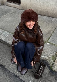 Γυναίκα με καφέ γούνινο καπέλο και δερμάτινο παλτό καθισμένη στο πεζοδρόμιο, χαμογελαστή, με σκούρα τζιν, παπούτσια με τετράγωνη μύτη και μία μικρή καφέ τσάντα δίπλα της.