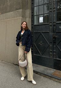 Denim jacket, green knitted top, beige wide-leg trousers, and a white patterned handbag; white trainers with beige accents. Urban backdrop.