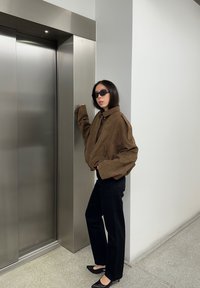 Veste bomber marron en daim avec manches surdimensionnées, pantalon noir cintré et ballerines noires pointues, debout à côté d'un ascenseur métallique.