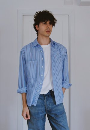 Camisa de botones a rayas azul y blanca, con dos bolsillos delanteros, mangas enrolladas, combinada con jeans azules. Fondo neutro.