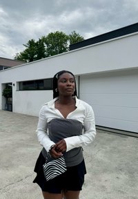 Weißes Langarmhemd mit grauem Oberteil, kombiniert mit einem schwarzen Rock. Hält eine kleine schwarz-weiß gestreifte Handtasche. Betonhintergrund.