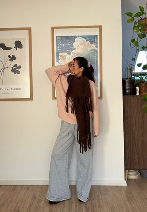 Mujer de pie en el interior vistiendo un suéter rosa claro, pantalones anchos grises, bufanda larga marrón, con obras de arte enmarcadas y plantas en el fondo.