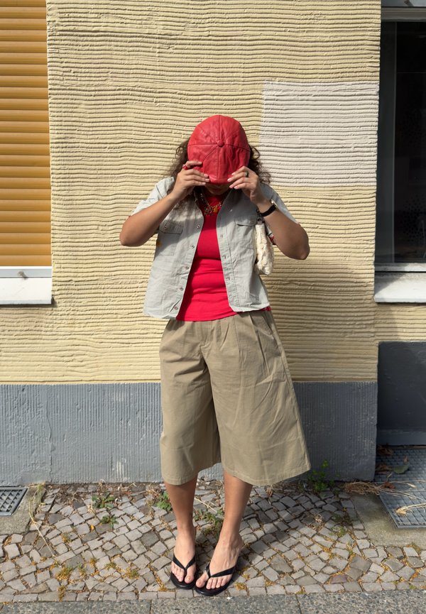 Persoon die een rode pet rechtzet, draagt een rood overhemd, een beige jas, een wijde beige broek en zwarte sandalen, staand op een kasseienpad tegen een structuurmuur.