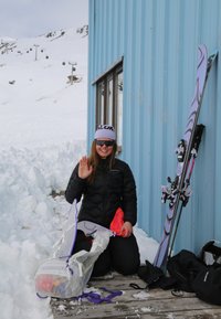 Personne vêtue d'un équipement d'hiver faisant signe de la main en s'agenouillant près d'un bâtiment bleu, à côté de skis et d'un sac à dos partiellement ouvert dans un cadre montagneux enneigé.