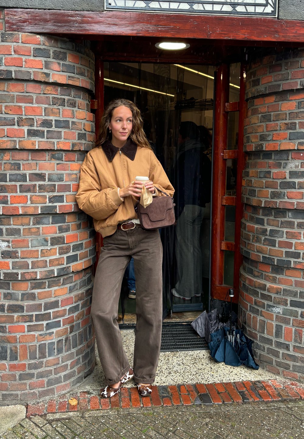 Giubbotto bomber tan con colletto in pelliccia sintetica marrone, pantaloni marroni, scarpe a pois, che tiene una tazza di caffè, appoggiato a un ingresso con parete di mattoni.
