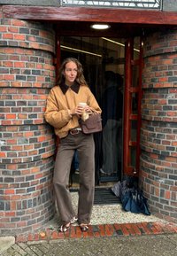 Giubbotto bomber tan con colletto in pelliccia sintetica marrone, pantaloni marroni, scarpe a pois, che tiene una tazza di caffè, appoggiato a un ingresso con parete di mattoni.