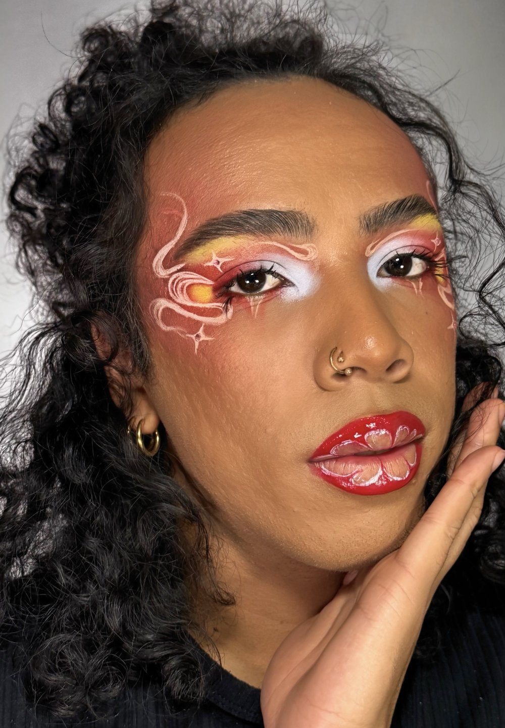 Närbild av ett ansikte med intrikat smink. Huden har en ljusbrun ton; djärva röda läppar med blommönster. Bakgrunden är mjuk grå.