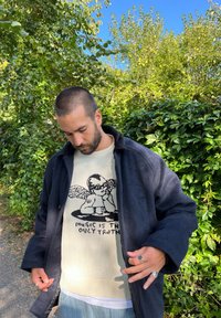 Casaco de lã azul-marinho sobre um suéter creme com uma gráfica de uma figura alada e o texto "A MÚSICA É A ÚNICA VERDADE." Calças de denim claras.