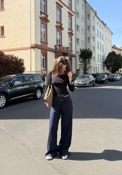 Mujer con camiseta negra de manga larga y pantalones anchos está en una calle soleada con coches aparcados y edificios residenciales de varios pisos.