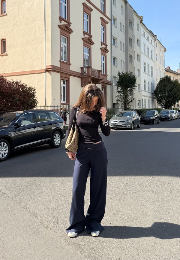 Žena u crnoj bluzi dugih rukava i širokim hlačama stoji na sunčanoj ulici s parkiranim automobilima i stambenim višekatnicama.