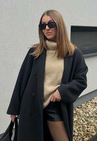 Femme portant des lunettes de soleil noires, un manteau sombre, un pull à col roulé côtelé beige, une jupe en cuir noire, tenant un sac noir, debout près d'un mur blanc avec des pierres.