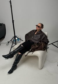 Jachetă din piele maro, jeansi negri rupți, cizme negre până la genunchi, ochelari de soare negri, stând pe un scaun alb curbat cu o geantă marron.