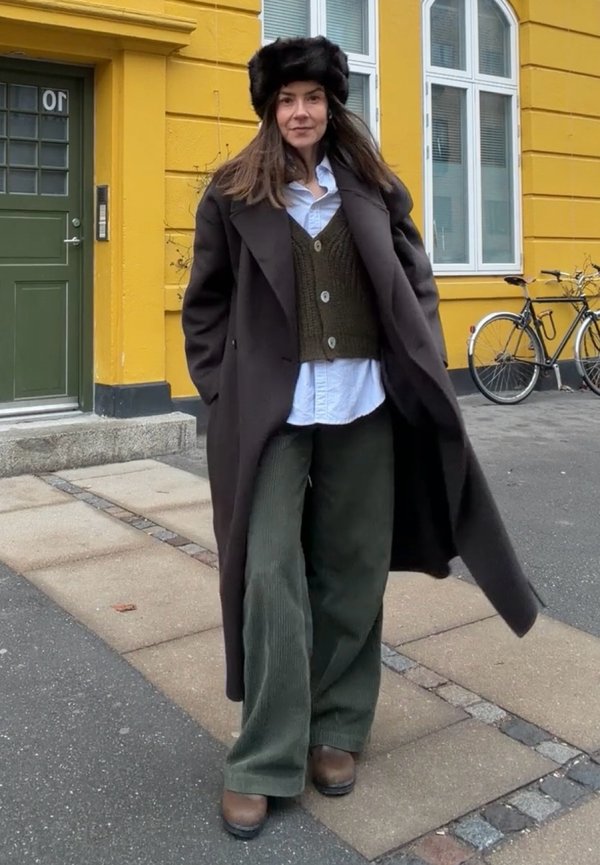 Femme portant un long manteau sombre, un pantalon vert à jambes larges, des chaussures marron, un gilet en tricot, une chemise bleu clair et un chapeau en fourrure noir, debout sur le trottoir près d'un bâtiment jaune.