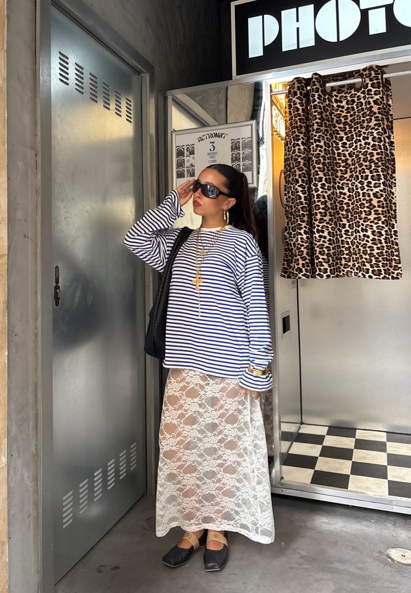 Mujer con camiseta de manga larga a rayas, falda blanca de encaje y gafas de sol, de pie cerca de una puerta metálica junto a una cabina de fotos con cortina de estampado de leopardo.
