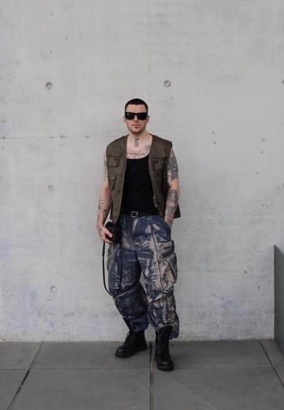 Chaleco cargo tan sobre una camiseta negra de tirantes, combinado con pantalones cargo azules y beige de estampado y botas negras con cordones. Los accesorios incluyen gafas de sol y una cámara.