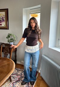 Joven con cabello largo y ondulado, lleva una blusa negra, camisa de encaje blanca, jeans azules y zapatos oscuros, de pie en interiores junto a una ventana y muebles de madera.