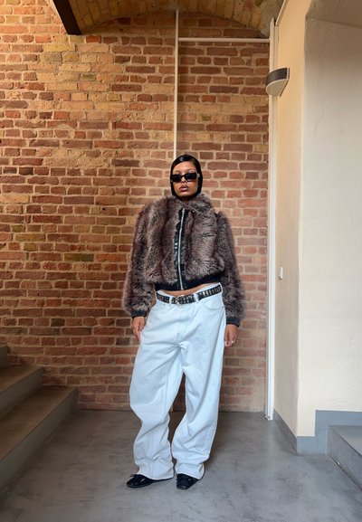 Chaqueta de piel sintética marrón con ribete negro, diseño corto, llevada sobre una blusa negra, combinada con pantalones anchos blancos y zapatos negros.