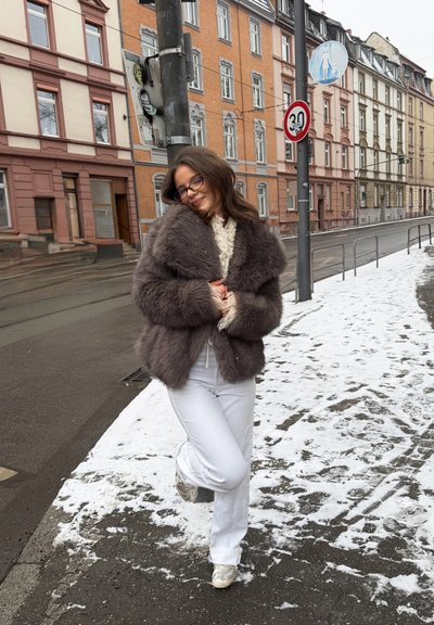 Mujer con gafas que lleva un abrigo de piel marrón, pantalones blancos y zapatillas, de pie en una acera nevada con edificios urbanos y señales de tráfico detrás de ella.