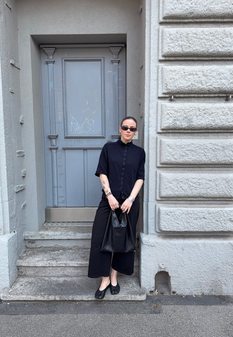 Woman in all-black outfit and sunglasses stands on stone steps holding a black bag in front of a grey ornate door and textured wall.