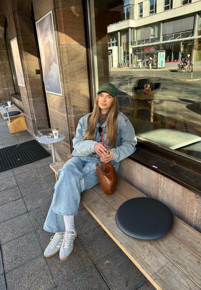 Chaqueta de denim, camiseta gris y jeans de color azul claro. Bolso marrón al lado. Zapatillas grises con calcetines blancos. Banco de madera y objeto negro circular cerca.