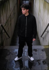 Black pinstriped zip-up jacket over a black shirt, teamed with loose black trousers and white trainers, standing on wet pavement in a narrow corridor.