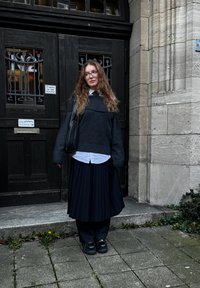 Γκρι oversized πουλόβερ τοποθετημένο πάνω από λευκό πουκάμισο με γιακά, συνδυασμένο με μαύρη πλισέ φούστα. Μαύρα παπούτσια και μια υφασμάτινη τσάντα ώμου.