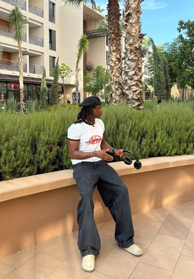 Pantalones de mezclilla negros acampanados, camiseta gráfica blanca, zapatos beiges. Sosteniendo auriculares negros, sentado en un banco de piedra claro entre la vegetación.