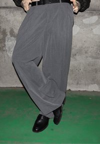 Oseba v sivih hlačah širokih nog z navpičnimi črtami, črnim pasom in črnimi usnjenimi čevlji, stoji na zeleni tleh ob teksturirani sivi steni.