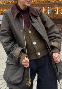 Bruine waterdichte jas met een bruine kraag, gouden rits en grote zakken. Draagt een groene gebreide trui en donkere broek eronder.