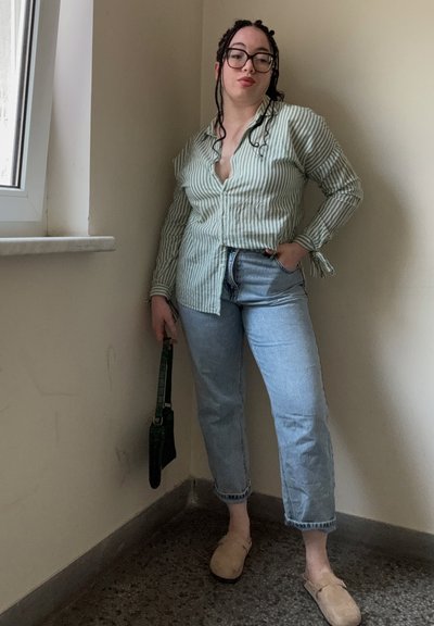 Camisa de botones a rayas verdes y blancas, jeans de mezclilla azul claros, zapatos beige de tipo slip-on y un pequeño bolso cruzado verde.