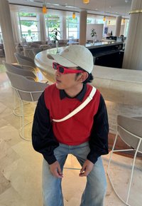 Red sleeveless sweater worn over a black shirt, matched with light blue jeans. Complemented by a beige cap and red sunglasses, seated in a contemporary lounge.