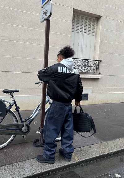 Chaqueta bomber negra con el texto "UNIFORM", sudadera con capucha gris, pantalones de mezclilla oscuros de gran tamaño, sosteniendo una bolsa negra, cerca de una bicicleta.