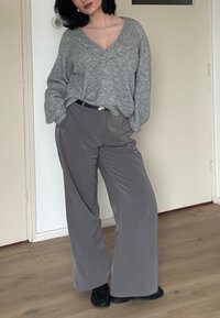 Pull gris en maille, coupe ample avec col en V ; pantalon gris à jambes larges ; chaussures noires ; sol en bois et murs clairs en arrière-plan.