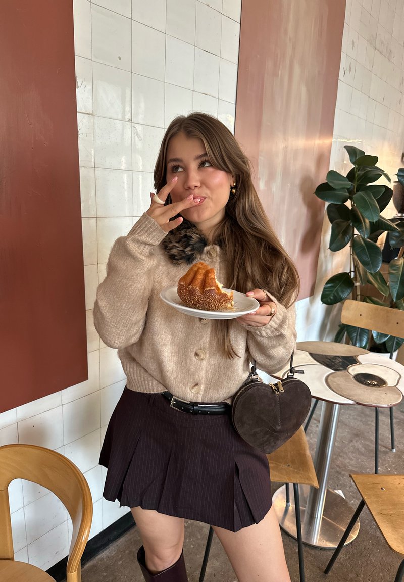 Beige strikket cardigan med knapper, pelsdetalj på kragen. Mørk plisséet skjørt, holder en tallerken med bakverk, tilbehør med en hjerteformet veske.
