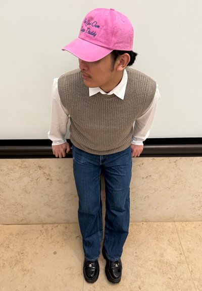 Gorra rosa con el texto "Sé tu propio Sugar Daddy", chaleco de punto marrón sobre una camisa blanca, vaqueros rectos azules y zapatos negros.
