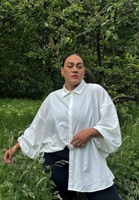 Weißes, übergroßes Hemd mit offenen Kragen, lockeren Ärmeln und strukturiertem Stoff; getragen mit schwarzen Leggings in einer natürlichen Umgebung.