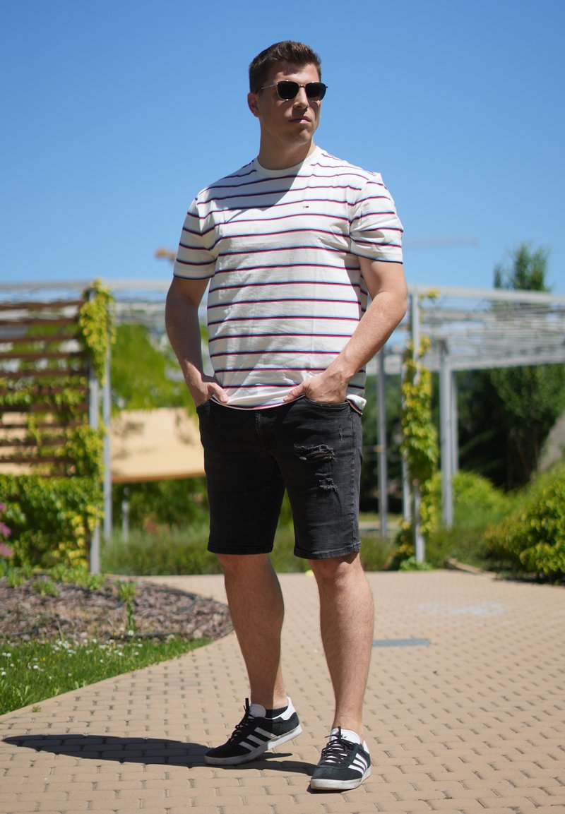 Mann mit Sonnenbrille, weiß gestreiftem T-Shirt, schwarzen Shorts und Turnschuhen steht draußen auf einem sonnigen gepflasterten Weg mit Grün im Hintergrund.