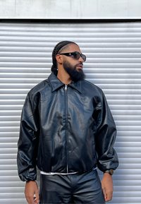 Czarna skórzana kurtka bomberka z zamkiem błyskawicznym, szerokim kołnierzem i długimi rękawami. Nosi się ją na białą koszulę i łączy z czarnymi skórzanymi spodniami.