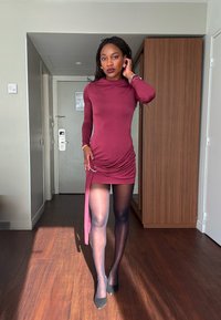 Robe longue bordeaux à manches longues avec un détail froncé sur le côté, présentant une fente haute. Portée avec des talons noirs et des collants noirs transparents. En arrière-plan, un mobilier en bois.