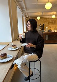 En person i en tykk, mørk brun høyhalset genser sitter på en svart krakk og holder en hvit kopp, med bakverk på tallerkener i nærheten, i en kafé.