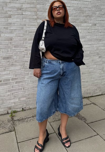 Persona con cabello largo rojizo, camiseta negra de manga larga, shorts anchos de mezclilla, sandalias negras, bolso blanco de hombro, de pie contra una pared de ladrillos gris.