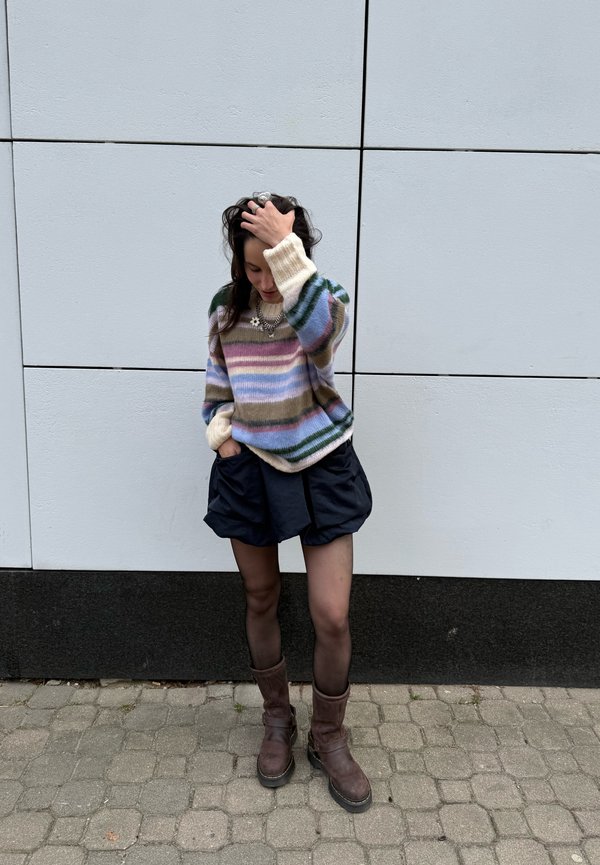 Joven mujer de pie contra una pared de azulejos, lleva un suéter de rayas, pantalones cortos negros, medias transparentes, botas marrones, con una mano tocándose el pelo.