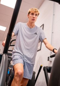 Tricou sportiv gri deschis cu dungi verticale și logo negru, purtat împreună cu șorturi asortate, de către o persoană pe o mașină de exerciții.