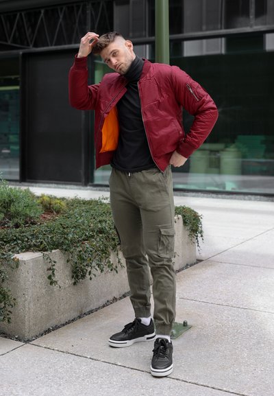 Chaqueta bomber roja con forro naranja, suéter de cuello alto negro, pantalones cargo oliva y zapatillas negras. El entorno presenta jardineras de concreto y arquitectura moderna.