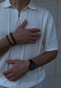 Polo bianco a maniche corte con zip a metà e righe verticali texturizzate. Gli accessori includono braccialetti neri di perline e un orologio da polso nero.