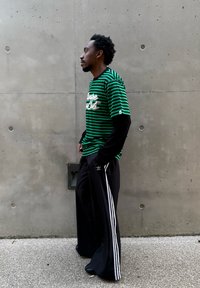 Zielona i czarna koszulka w paski z długimi czarnymi rękawami, zestawiona z czarnymi spodniami o szerokich nogawkach z białymi paskami po bokach, na szarym tle.