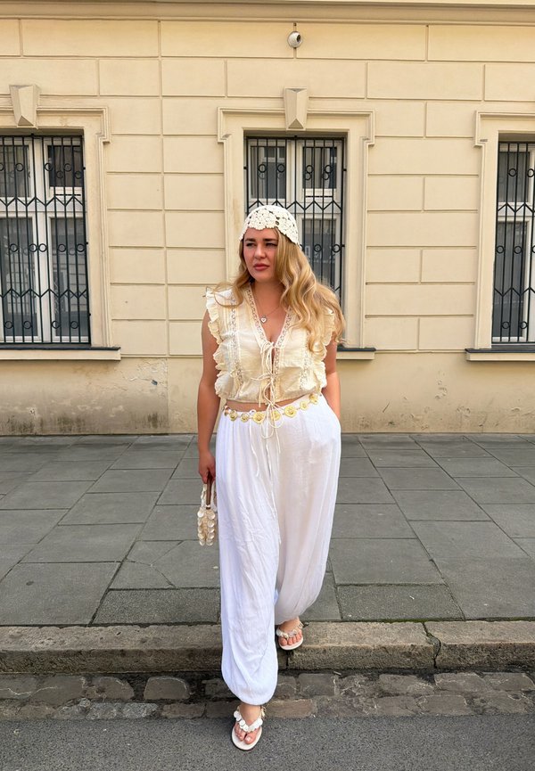 Kobieta z długimi blond włosami, ubrana w kremowy top z falbankami, białe spodnie, pasek z muszlami i kapelusz, spacerująca po miejskim chodniku w pobliżu beżowego budynku.